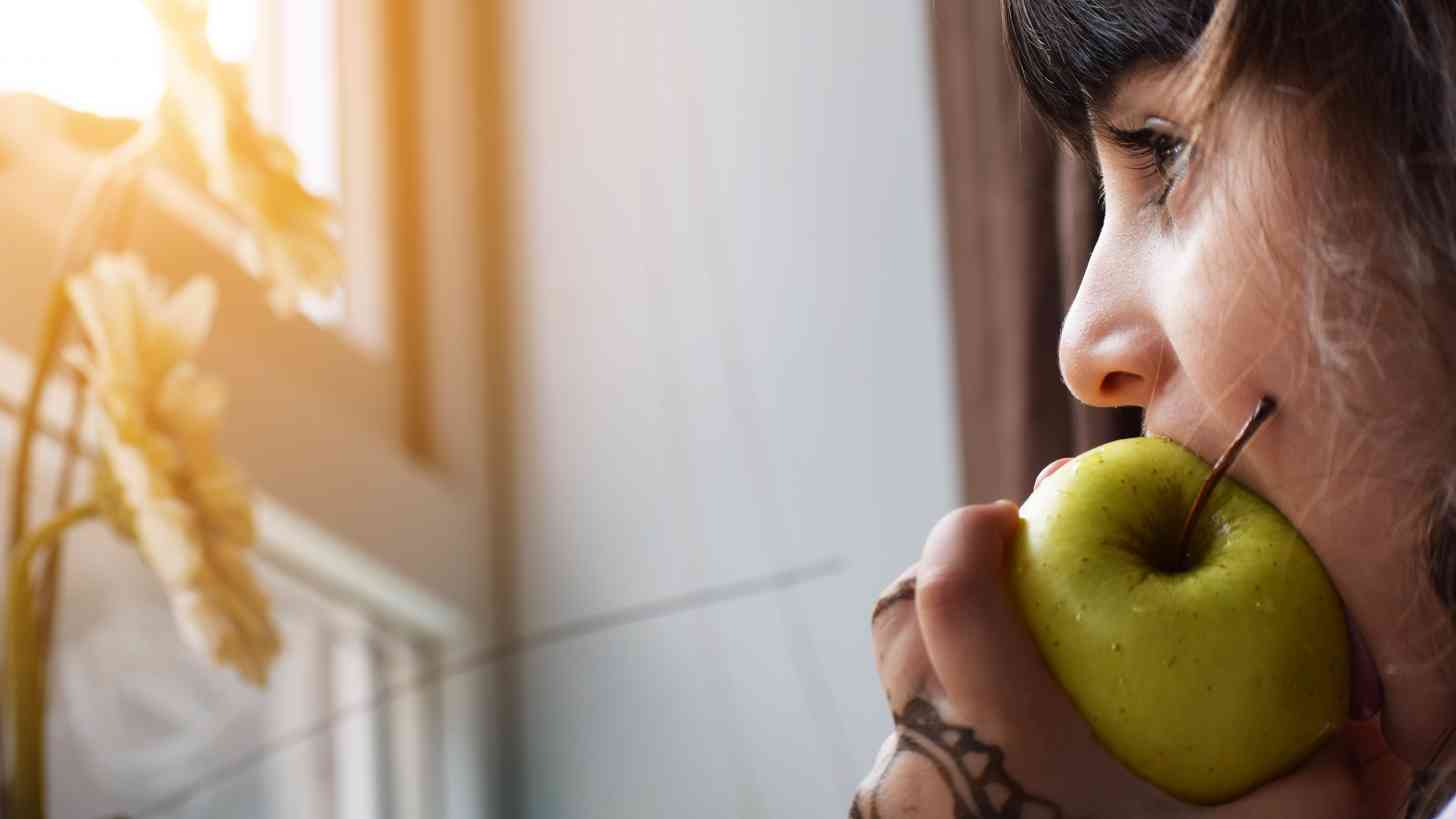 In 2019, a person holds a green apple near their mouth, gazing out a sunlit window. The serene scene contrasts sharply with the memory of a measles outbreak discussed by MDs. Meanwhile, flowers bloom gently in the background.