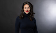 A global compliance manager with long dark hair and red lipstick is wearing a black turtleneck sweater, standing against a dark gray background, ready to address the team at Sermo.