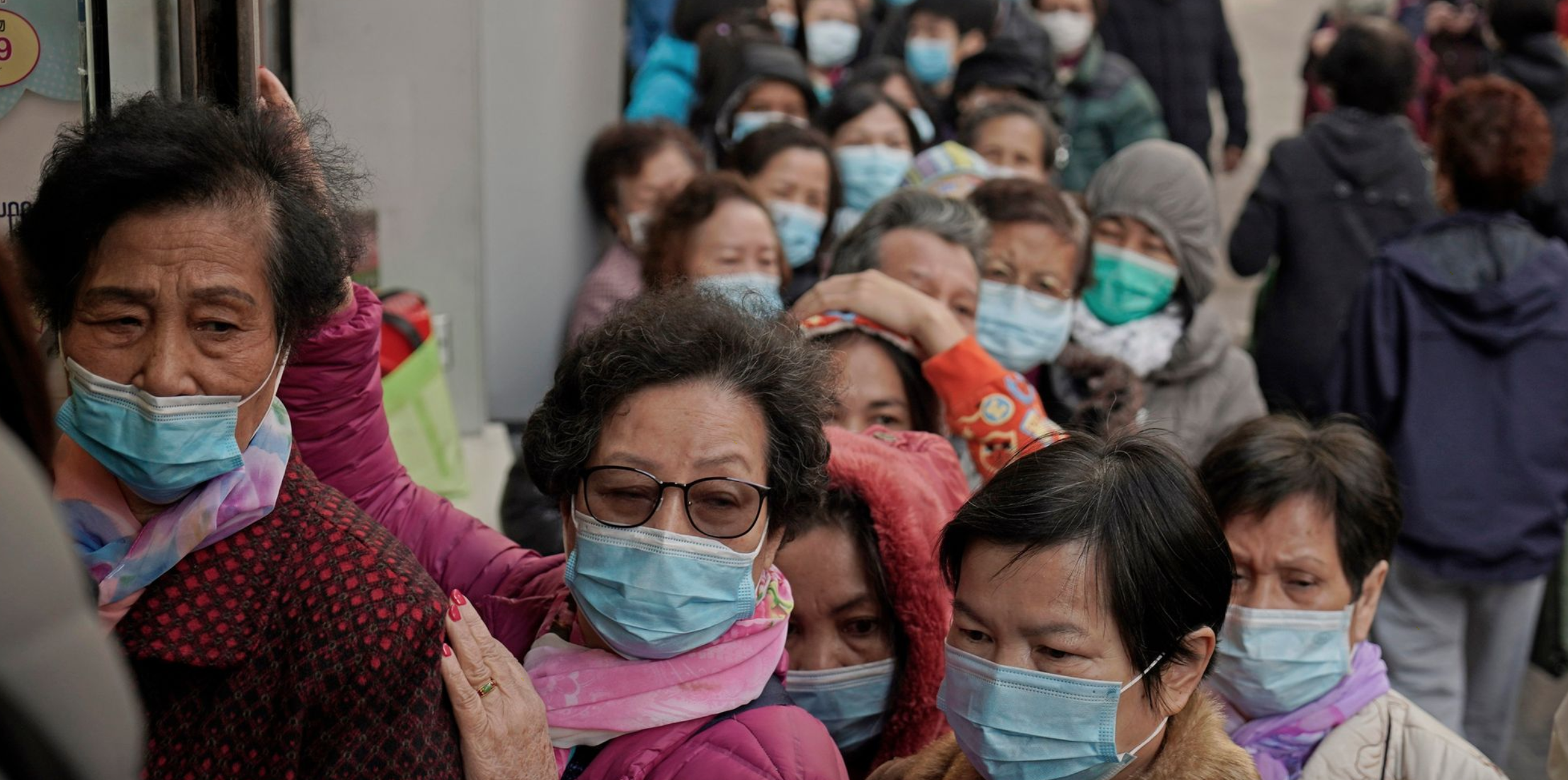 People wearing face masks stand closely together in a line, waiting outside a building, a scene reminiscent of the ongoing precautions due to the coronavirus.