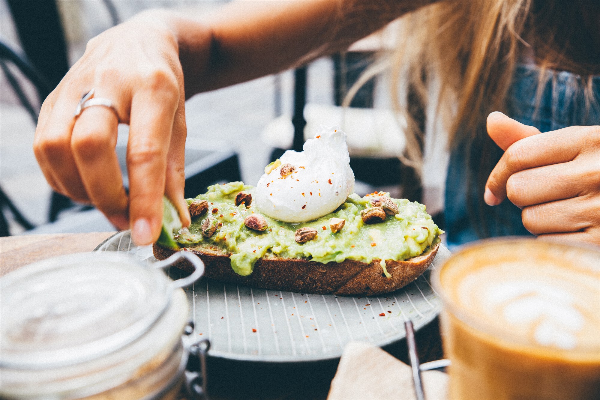 Figuring out the best way to eat avocado toast, a person savors each bite topped with a poached egg and nuts, alongside a steaming coffee.