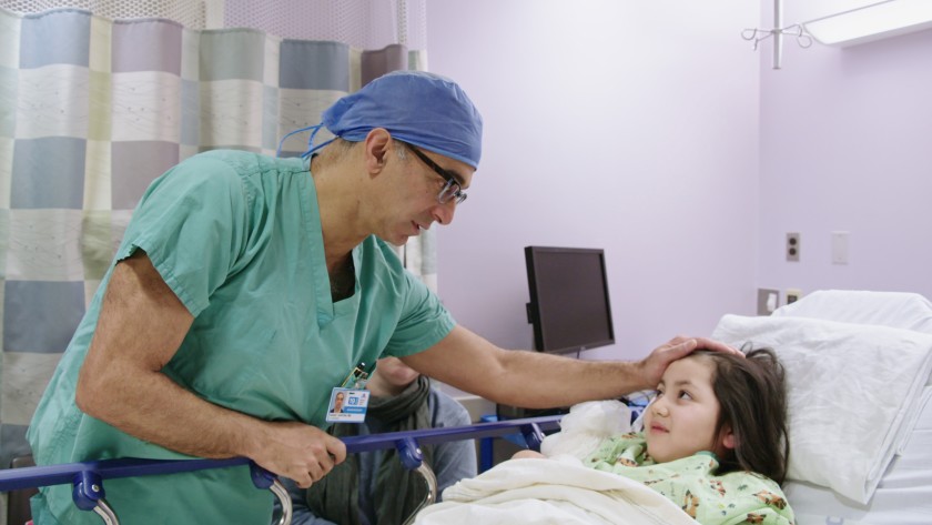 A medical professional in scrubs and a cap comforts a child lying in a hospital bed, highlighting the compassion often missing in our broken healthcare system.