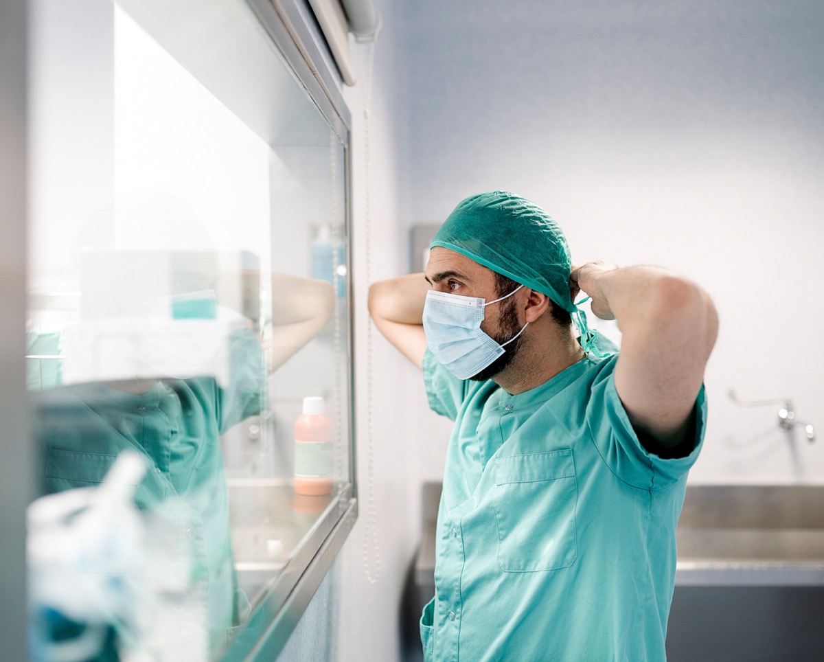 A healthcare worker in scrubs and a mask adjusts a surgical cap while looking in a mirror.