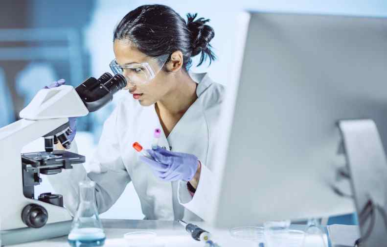 A scientist in a lab coat examines a test tube under the microscope, exploring hydroxychloroquine as a potential coronavirus treatment. A computer and various lab equipment fill the table, suggesting involvement in a global survey of medicinal efficacy.