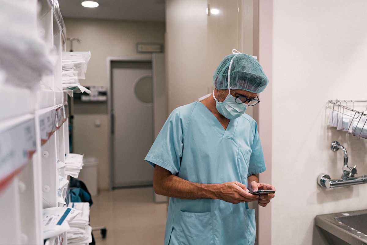 A medical professional in scrubs and a surgical cap stands in a hospital room, seeking a doctor's opinion via smartphone.