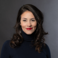 A woman with long dark hair, wearing a black turtleneck sweater, smiles against a dark gray background.