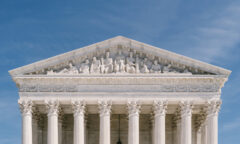 Front view of the U.S. Supreme Court building with its majestic columns supporting intricate sculptures on the facade, all under a clear blue sky.