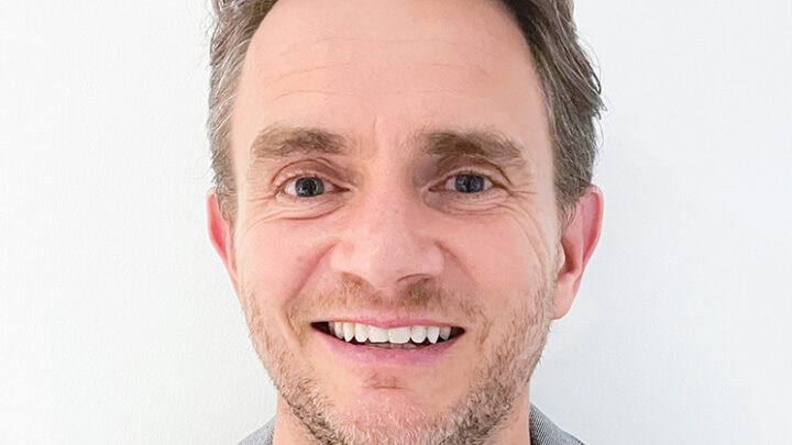 Smiling person with short, styled hair, wearing a grey blazer and white shirt, against a plain white background.