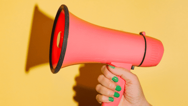 A pink megaphone, symbolizing innovative marketing strategies, is held by a hand with green-painted nails against a vibrant yellow background.