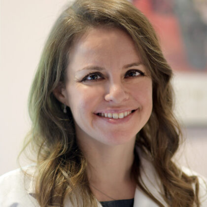 Eleonora Teplinsky, MD, a smiling person with long, wavy hair, stands indoors wearing a white coat.