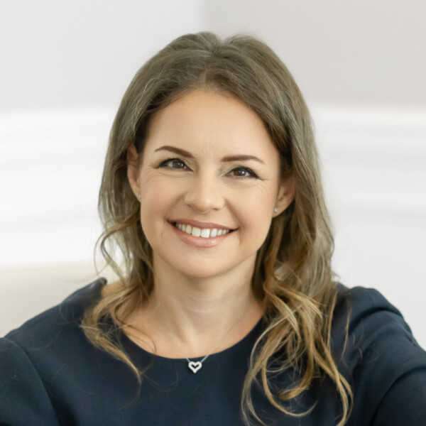 Eleonora Teplinsky, MD, a woman with long brown hair in a dark short-sleeve dress, sits smiling at a wooden table in a bright, minimalist office.