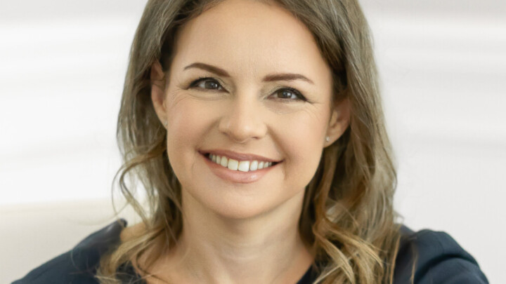 Eleonora Teplinsky, MD, a woman with long brown hair in a dark short-sleeve dress, sits smiling at a wooden table in a bright, minimalist office.