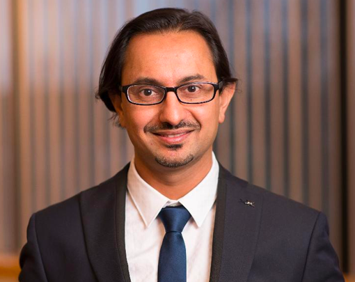 Una persona in abito nero e cravatta, con occhiali, sta in piedi davanti a uno sfondo a strisce. L'individuo composto trasuda professionalità, forse incarnando la competenza di qualcuno come Shivang Joshi, MPH.