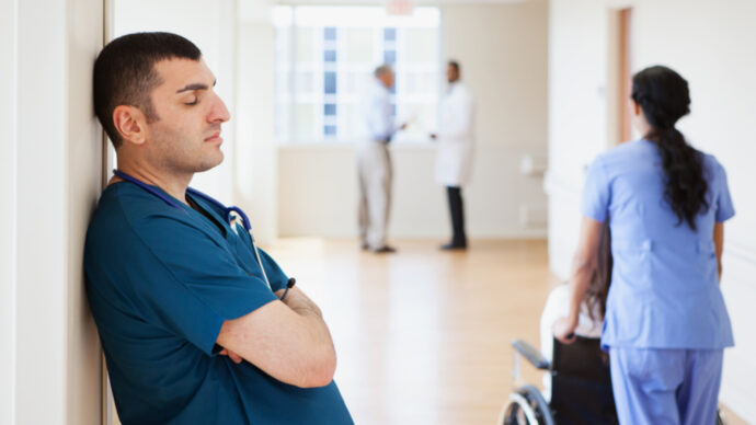 doctor leaning against a wall in a hospital hallway, eyes closed, arms crossed