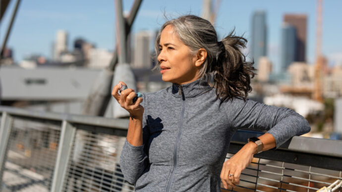 A woman with gray hair in a ponytail leans on a railing, holding a hand exercise device, reflecting on recent HCP insights. She wears a gray zip-up jacket as the city skyline stretches behind her.