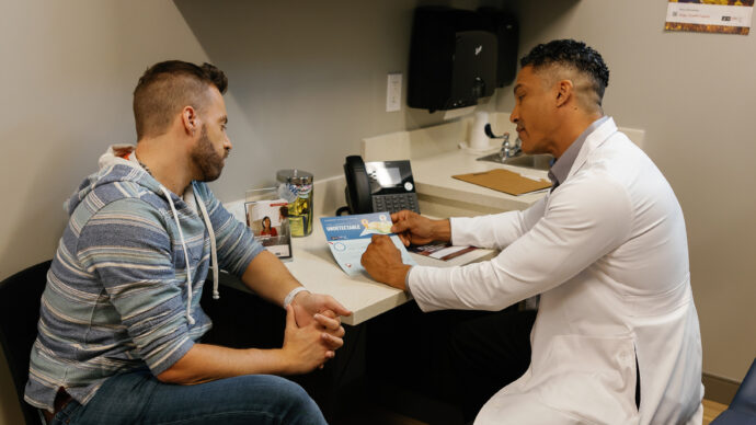 A doctor in a white coat discusses an informational pamphlet on HIV treatment with a seated patient in an office setting.