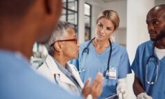 Medical professionals in scrubs have a discussion in a hospital setting. One person gestures while others, including experienced physicians and NPs, listen attentively, wearing stethoscopes and ID badges. The focus shifts to the ongoing "ill-trained NPs" controversy affecting team dynamics.