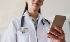 A person in a white coat with a stethoscope around their neck is holding a smartphone, possibly checking the latest updates on the 2024 presidential election.