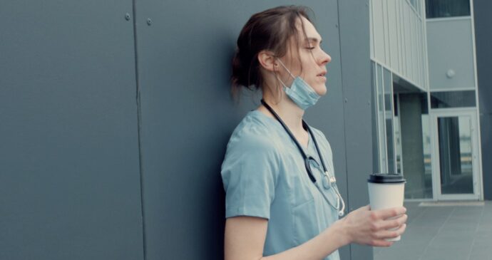A healthcare worker in scrubs and a stethoscope leans against a wall, holding a coffee cup, with a mask pulled below their chin.