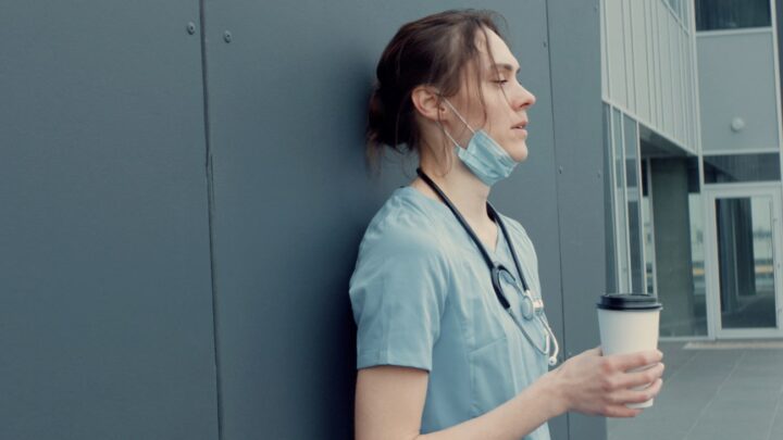 A healthcare worker in scrubs and a stethoscope leans against a wall, holding a coffee cup, with a mask pulled below their chin.