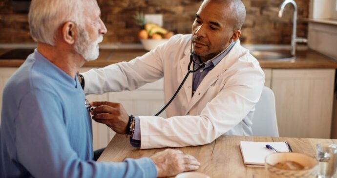 A doctor uses a stethoscope to examine an elderly man in a casual setting, with a notebook and a plant in the background, ensuring patient satisfaction.