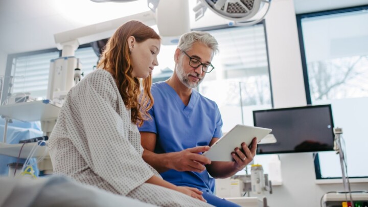 A physician in scrubs shows a patient in a hospital gown important medical information on a tablet, addressing the patient’s medical needs in a modern examination room.