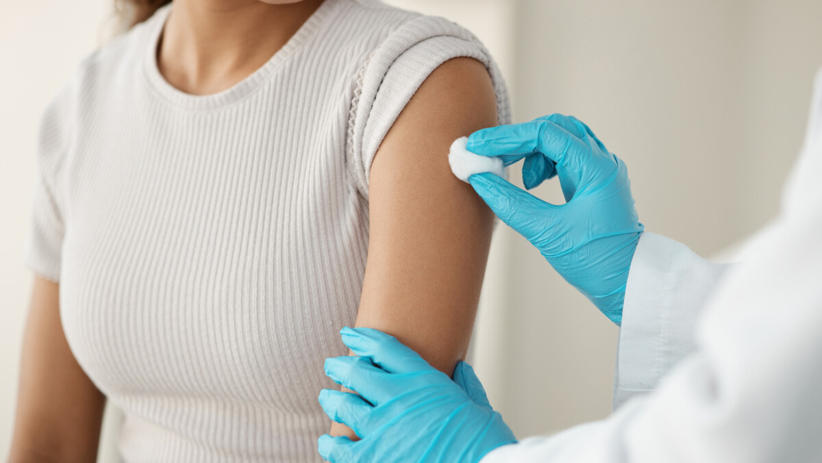 A healthcare provider wearing blue gloves cleans a woman's upper arm with a cotton ball in preparation for an injection, following the latest recommendations and ACIP guidelines.
