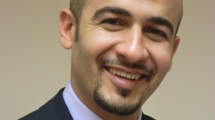 Amer Harky, FRCS, a man in a suit and tie, smiles at the camera against a plain beige background.
