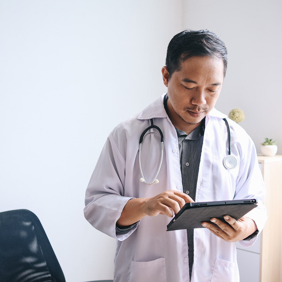 A doctor wearing a lab coat and stethoscope reviews information on a digital tablet in a well-lit office, exploring various medical specialities.