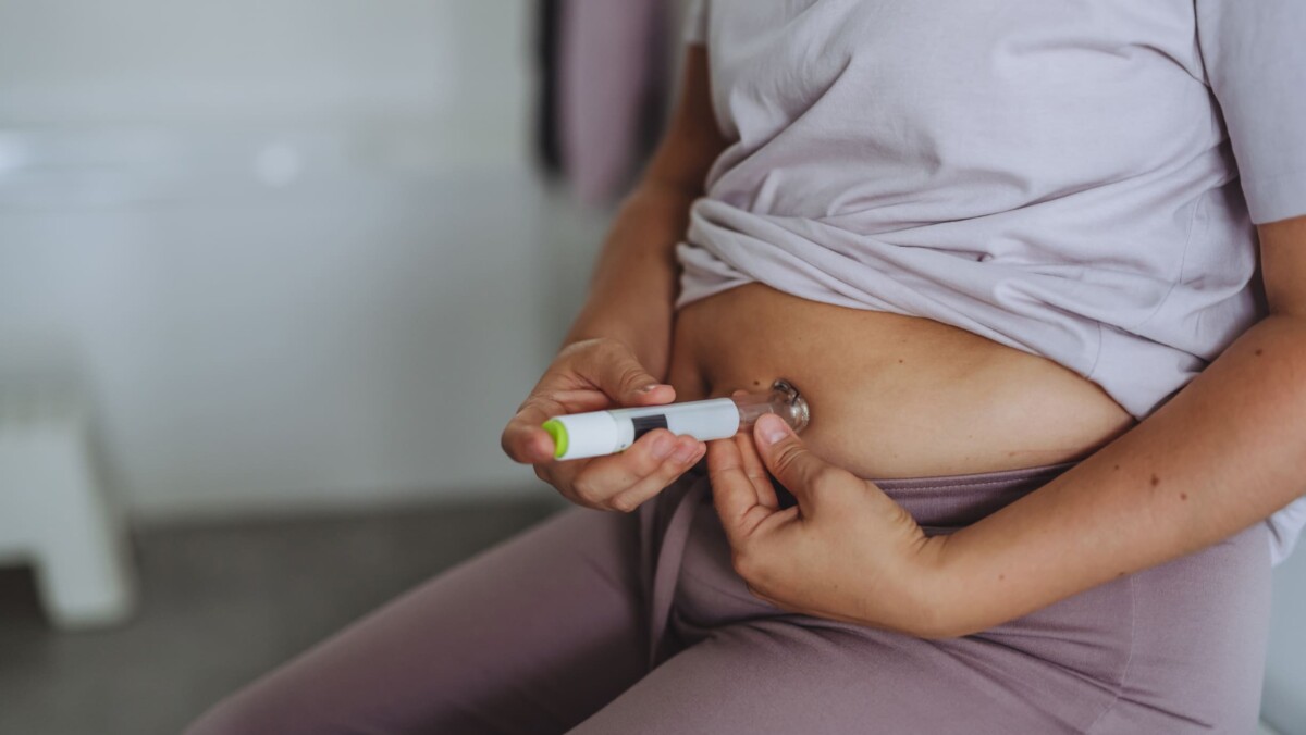 A person injects medication into their abdomen using an auto-injector pen while sitting down, with their shirt slightly lifted.