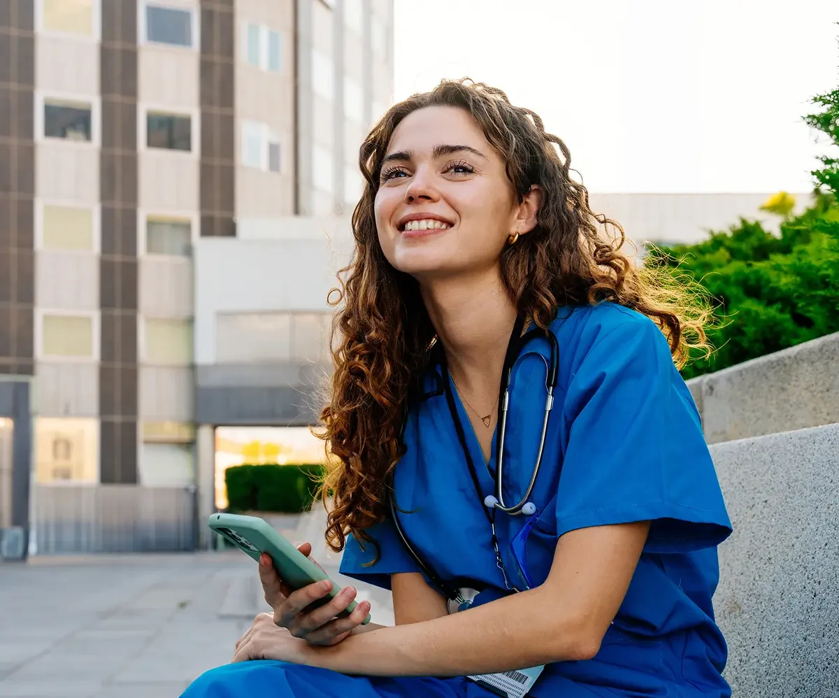 A healthcare worker in blue scrubs, representing diverse medical specialities, sits outdoors on a concrete bench, holding a phone and smiling, with a city building in the background.