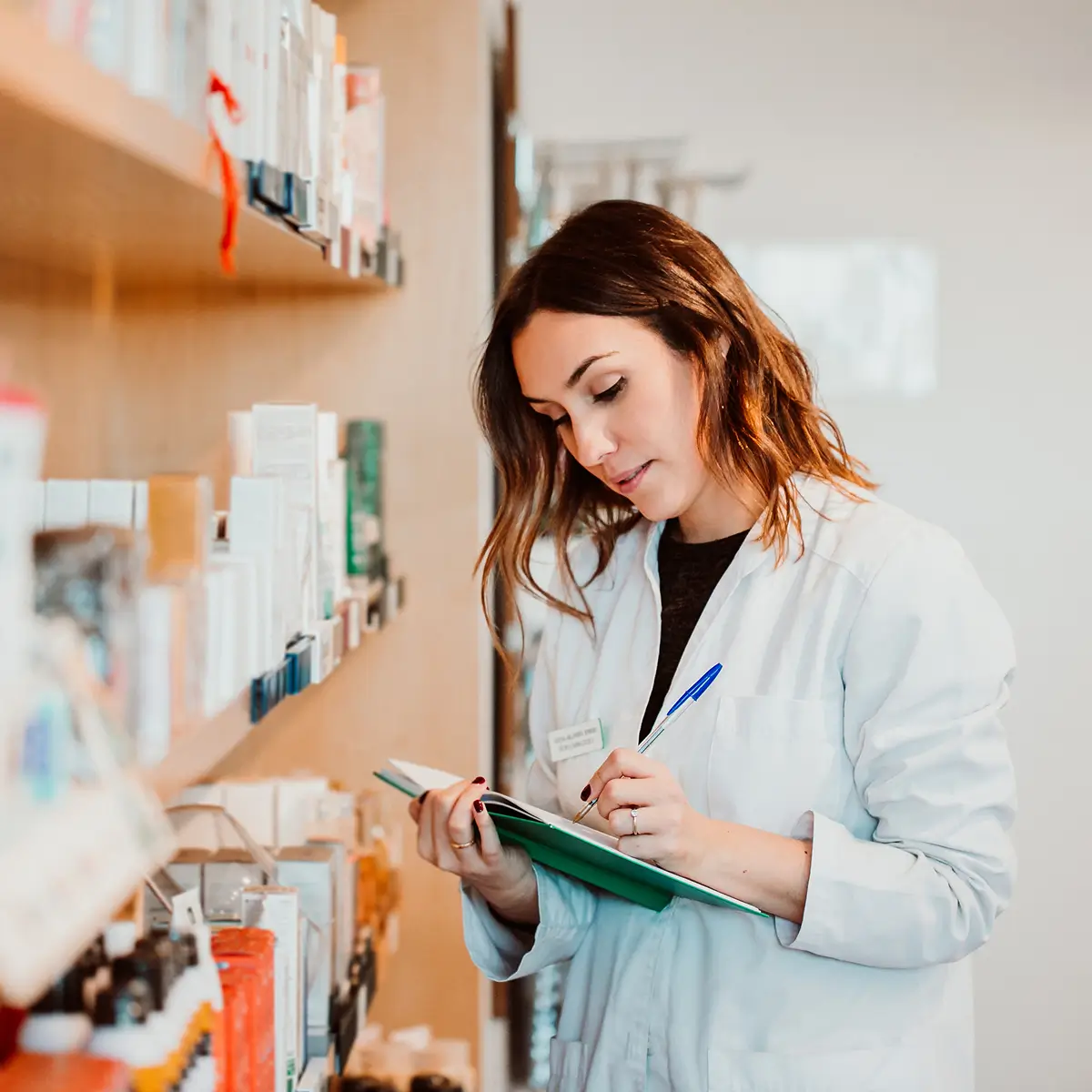 A woman in a white lab coat writes on a clipboard while standing next to shelves filled with various pharmaceutical products, reflecting her expertise in medical specialities.