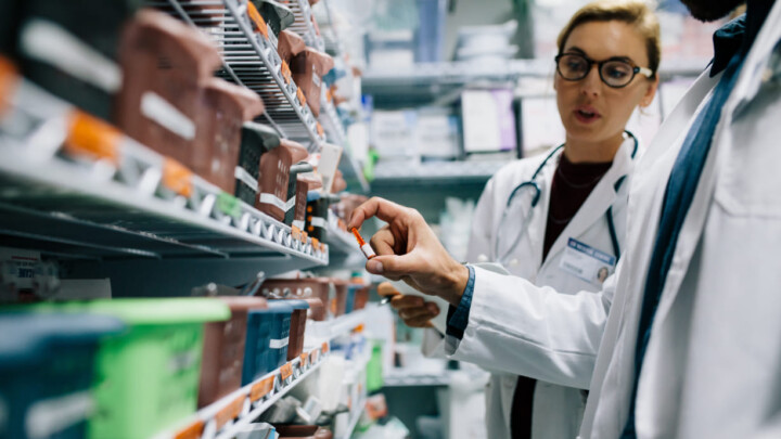 Two healthcare professionals in lab coats examine medication on shelves in a pharmacy or medical supply room, discussing the benefits of the TrumpRx discount card for physicians and patients.