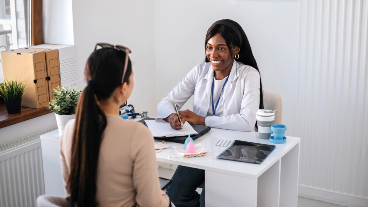 A healthcare professional sits at a desk, smiling and talking with a patient; contraceptive devices, a notepad, and results from medical market research surveys are on the table.
