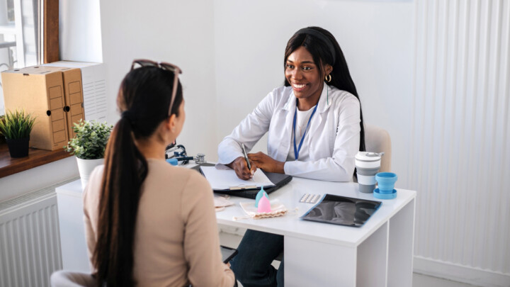 A healthcare professional sits at a desk, smiling and talking with a patient; contraceptive devices, a notepad, and results from medical market research surveys are on the table.
