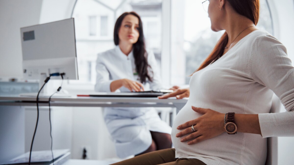 A pregnant woman sits in a doctor's office, holding her belly, while a healthcare professional speaks with her across a desk.