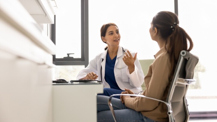A doctor sits at a desk speaking with a patient, who is seated across from her in an office setting, highlighting the impact of physician influence on consumer health decisions.