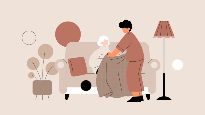 An elderly person sits on a sofa while another stands, placing a blanket over them—illustrating both the pros and cons of home health nursing; a lamp and plants are nearby.