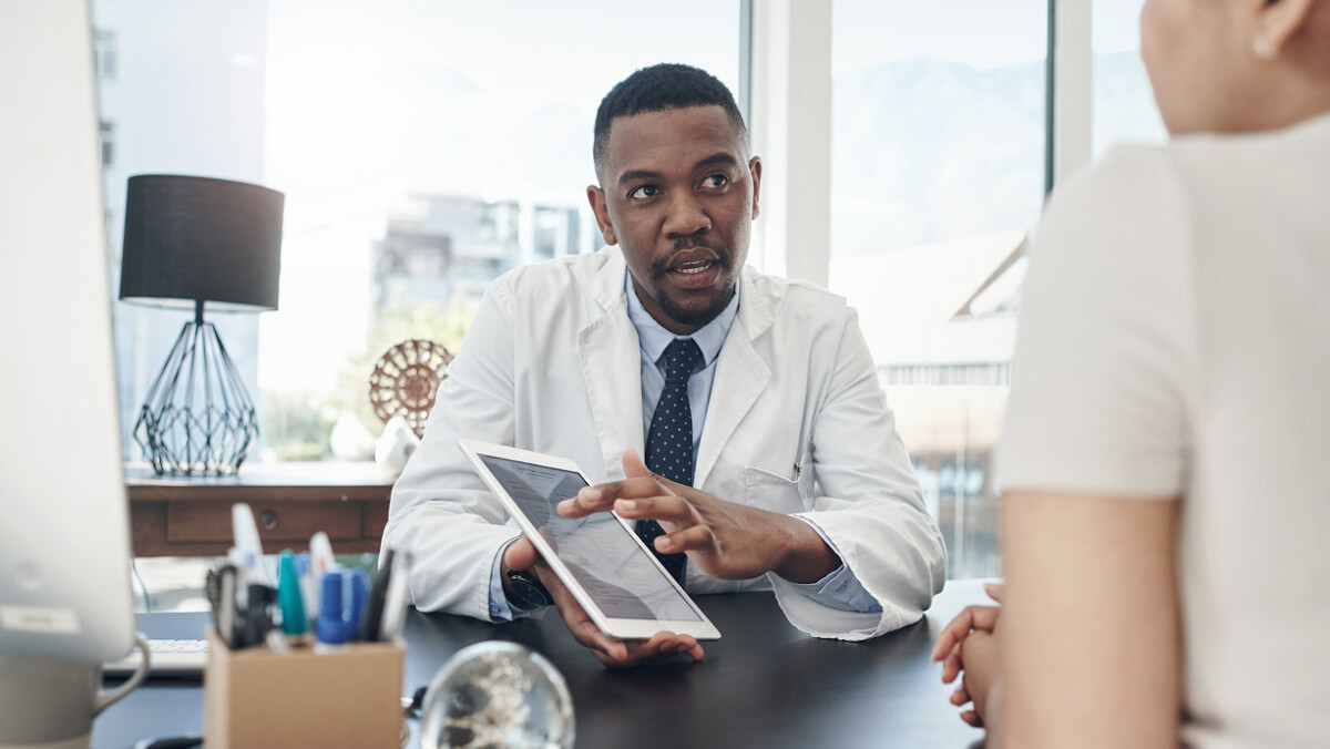 A doctor in a white coat sits at a desk, holding a tablet and explaining something to a patient across from him in a bright office, illustrating how technology adoption is transforming the way physicians engage with patients.