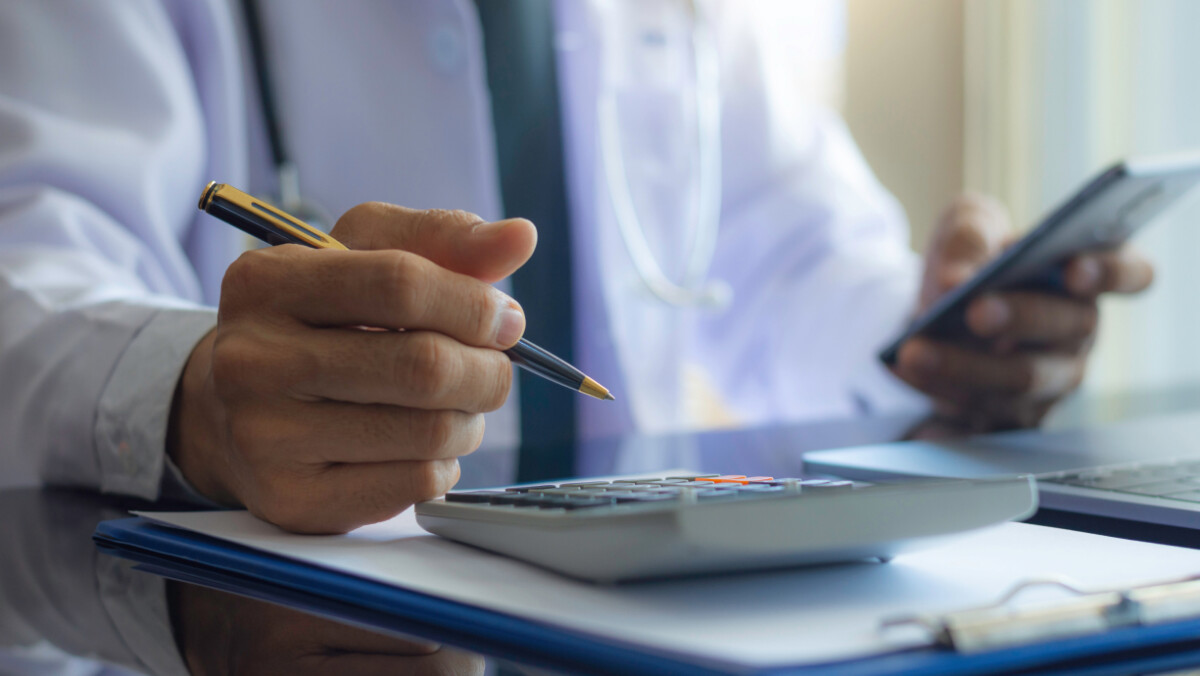 A person wearing a white coat uses a calculator and holds a smartphone, with a clipboard and laptop on the desk—illustrating the basics of finance & investing for physicians.