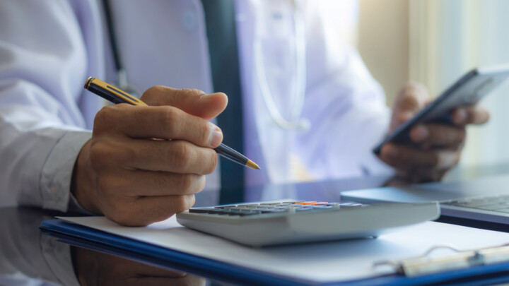 A person wearing a white coat uses a calculator and holds a smartphone, with a clipboard and laptop on the desk—illustrating the basics of finance & investing for physicians.