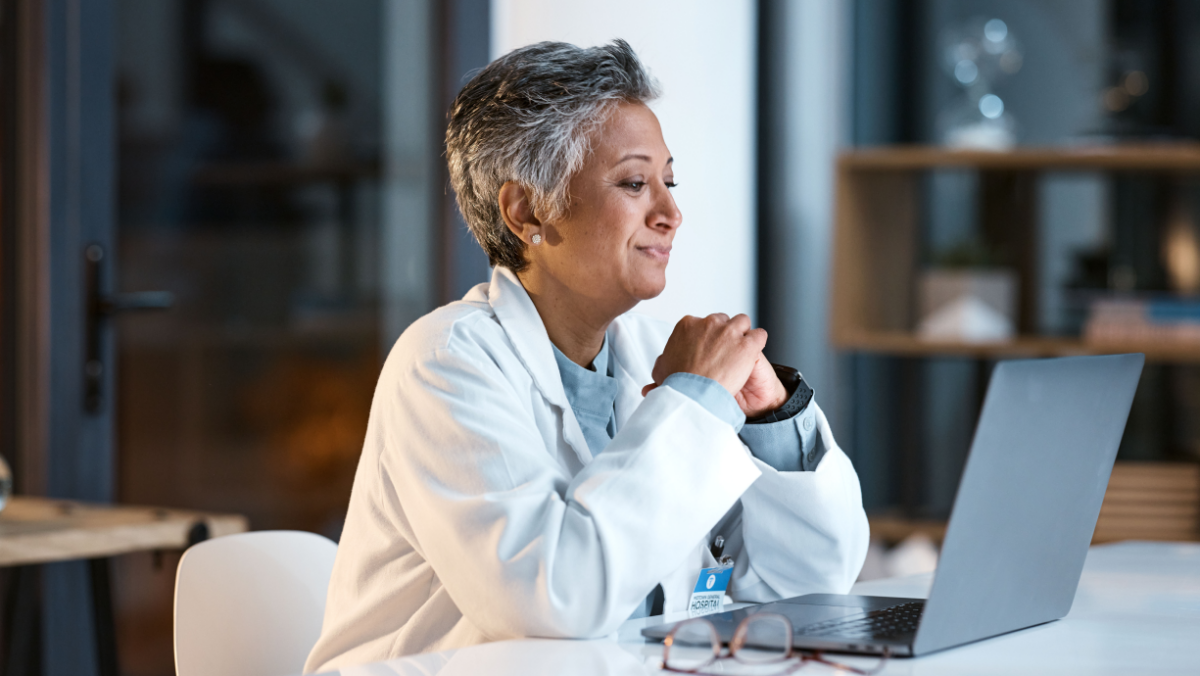 A doctor with short gray hair wearing a white coat sits at a desk looking at a laptop, appearing focused.