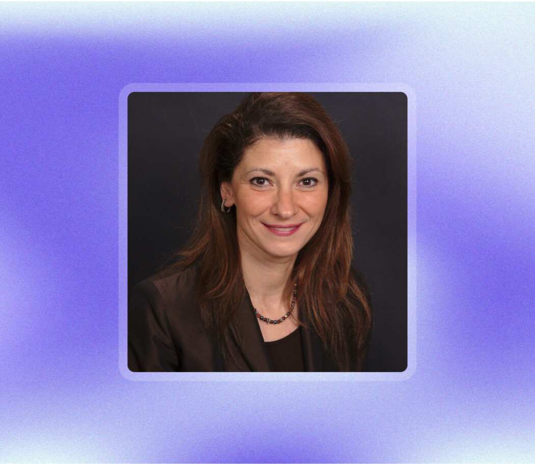 Woman with long brown hair in a dark blazer smiles at the camera, set against a black background with a purple gradient border, reflecting confidence and professionalism—qualities often associated with strong non-clinical skills.