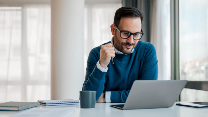 A man wearing glasses and a blue sweater sits at a desk with a notebook, cup, and laptop, appearing focused while looking at the screen in a bright office setting—perhaps researching non-clinical careers for physicians.