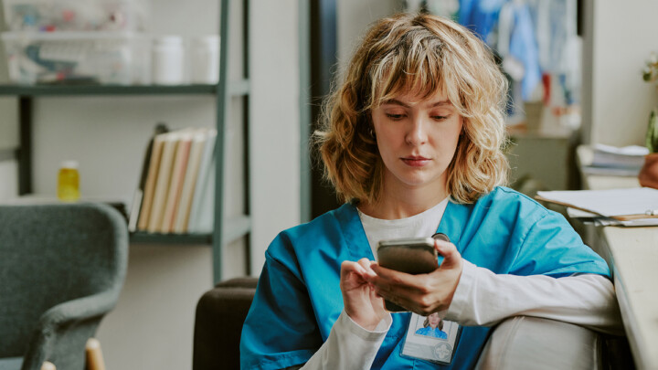 A person in blue medical scrubs sits indoors, looking at a smartphone—perhaps checking social media or reading about the latest developments in AI. Shelves with books and supplies are visible in the background.