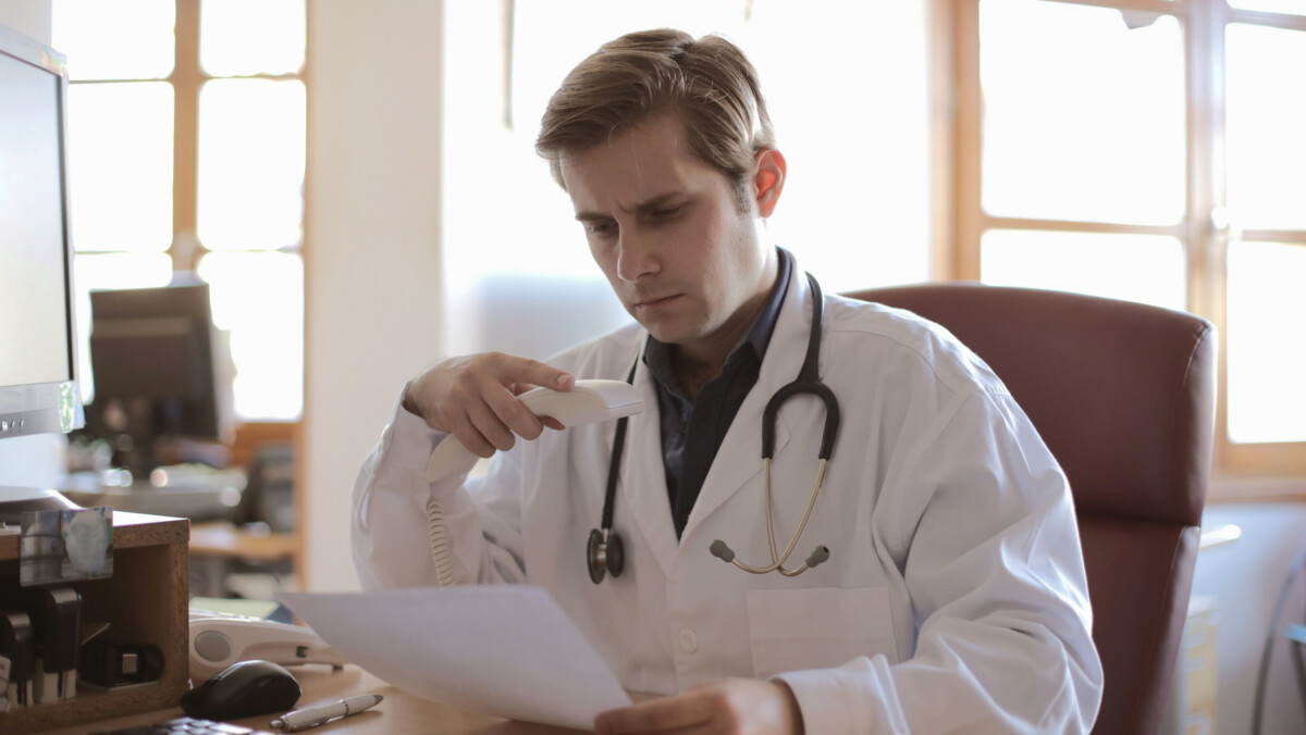 A doctor wearing a white coat and stethoscope sits at a desk, holding a phone and reading a document in a brightly lit office, carefully reviewing a document.