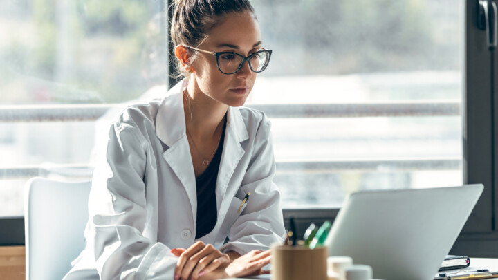 A doctor in a white coat sits at a desk looking at a laptop while reflecting on how to overcome feeling like you’re not enough as a doctor.