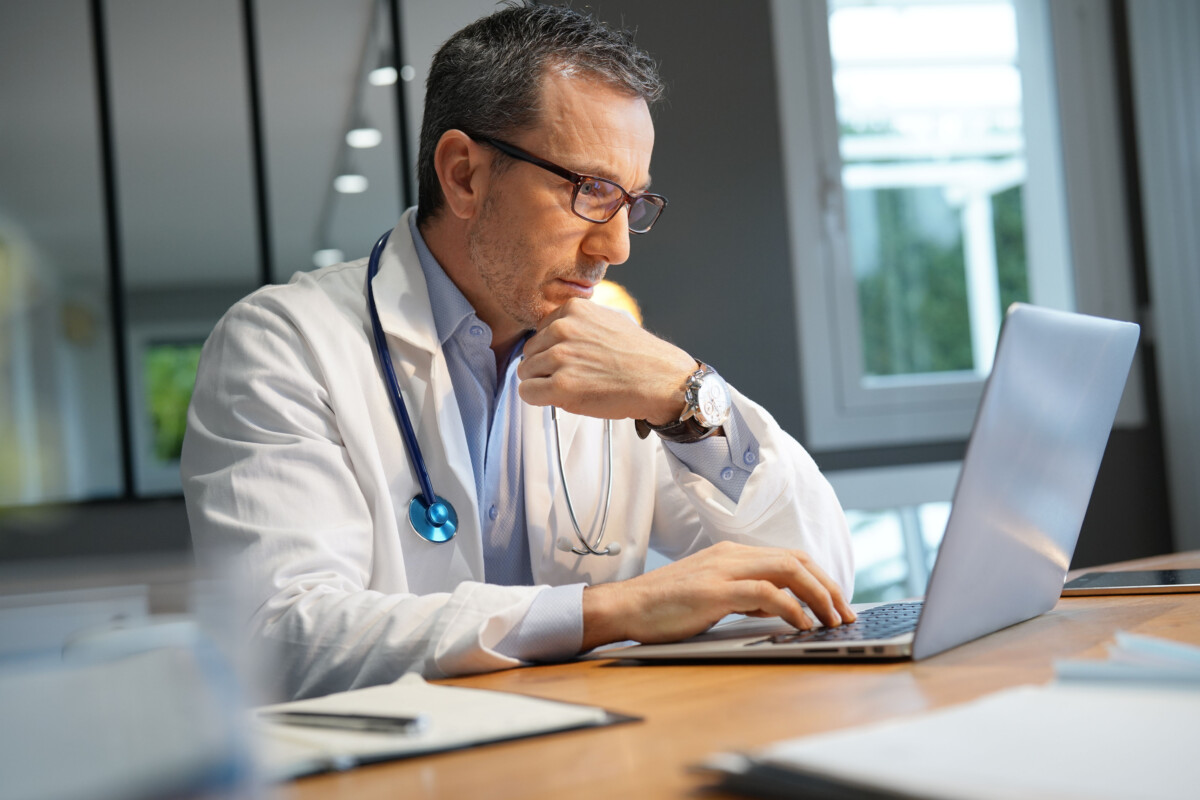 A doctor with a stethoscope around his neck sits at a desk and uses a laptop for AI verification in healthcare.