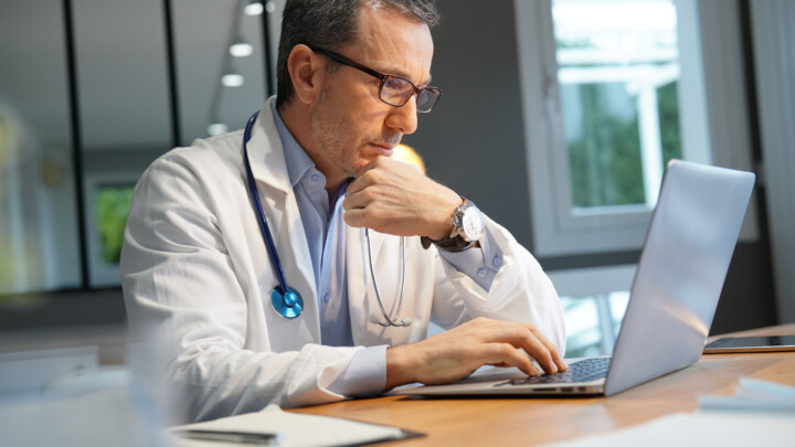 A doctor with a stethoscope around his neck sits at a desk and uses a laptop for AI verification in healthcare.