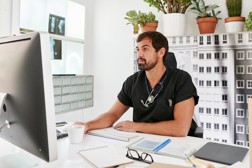 A clinician in black scrubs with a stethoscope around his neck sits at a desk, using healthcare technology to view a computer monitor.