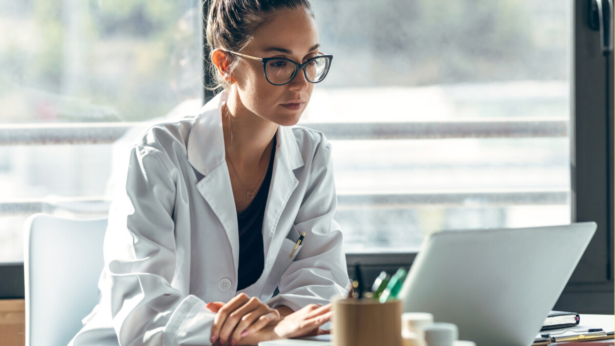 A physician in a white medical coat and glasses sits at a desk working on a laptop, illustrating the risks of online impersonation.